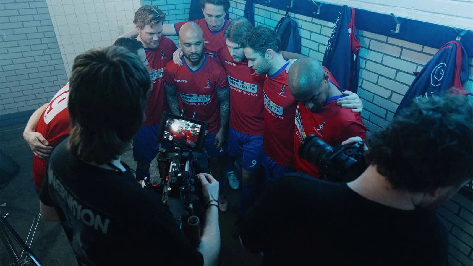 Voetballers in rode shirts staan in een kleedkamer in een kring, met twee cameramensen van Hokus Fokus op de voorgrond